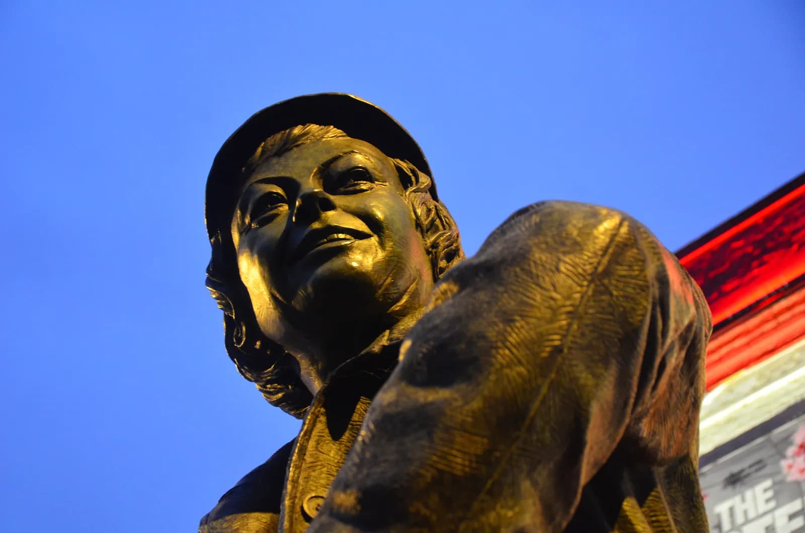 A low-angle shot captures a bronze statue of a person with a slight smile and tilted head, brightly lit under a vivid blue sky, with a red light fixture and a blurred sign partially visible in the upper right.